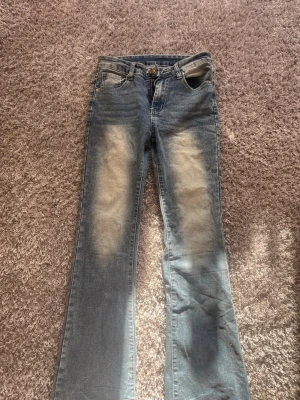 Blå bootcut jeans med slitningar - Snygga blå bootcut jeans med ljusa slitningar framtill och bakfickor med dekorativa detaljer. Klassisk femficksmodell med knapp och dragkedja. Perfekta för en avslappnad och trendig look.