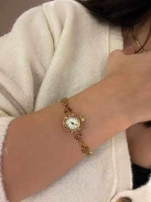 Guldig klocka med röda stenar - Snygg och elegant klocka med guldig boett och armband dekorerat med röda stenar. Klockan har en rund urtavla med vita detaljer och klassiska svarta siffror. Perfekt accessoar för att lyfta din outfit med lite extra bling.