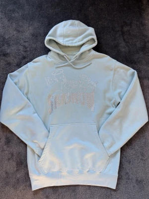 Ljusblå SKIDROW hoodie med strasstenar - Ljusblå hoodie från SKIDROW med huva och magficka. Framsidan har ett motiv av två änglar och texten SKIDROW i glittrande strasstenar. Mjuk bomullsmix och avslappnad passform, perfekt för en chill och trendig look.