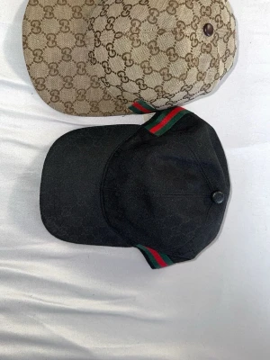 Gucci kepsar med GG-mönster - Två snygga kepsar från Gucci, en i beige med klassiskt GG-monogram och en i svart med subtilt GG-mönster. Båda har gröna och röda ränder på sidan och justerbar rem bak. Tillverkade i canvasmaterial med detaljer i bomull. Perfekt för dig som vill ha en exklusiv och trendig look.