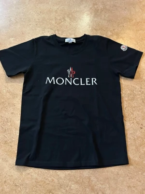 Svart Moncler t-shirt med logga - Snygg svart t-shirt från Moncler med stor vit och röd logga på bröstet och ett emblem på ärmen. Klassisk rund halsringning och korta ärmar. Tillverkad i mjuk bomull. Aldrig använd men lapparna är borttagna. Storlek small passar någon som är xs