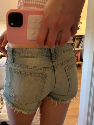 Ljusblå jeansshorts med fransig kant - Säljer ett par ljusblå denimshorts med fransig kant och klassiska fickor både fram och bak. Shortsen har låg midja och en avslappnad passform, perfekta för varma dagar. Snygg slitning på ena bakfickan för en cool look.