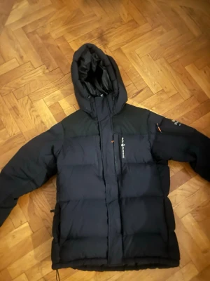 Svart dunjacka från Sail Racing DET finns ett problem med jackan och de kan man se på bild nummer 3  - Säljer en svart dunjacka från Sail Racing med huva och dragkedja framtill. Jackan har en puffig design och logga på bröstet. Perfekt för kalla vinterdagar tack vare det tjocka dunfodret. Snygg och funktionell med diskreta detaljer och fickor.