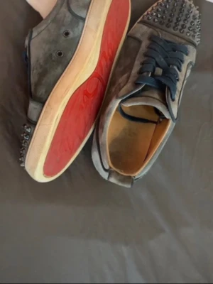 Gråa Louboutin sneakers med nitar - Unika gråa sneakers från Christian Louboutin i mocka med svarta nitar på tån och klassisk röd sula. Skorna har mörkblå snören och beige innersula. Perfekta för dig som vill sticka ut med en edgy och lyxig stil.