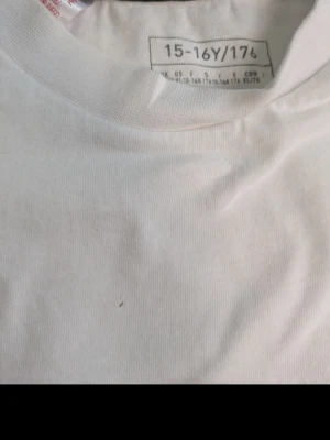 Vit Adidas t-shirt med logga - Säljer en klassisk vit t-shirt från Adidas med svart logga på bröstet. T-shirten har rund hals och är tillverkad i mjuk bomull, perfekt för en clean och sportig look. Enkel att matcha med jeans eller shorts.