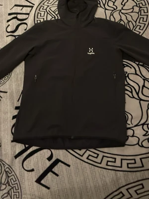 Svart softshelljacka från Haglöfs - Svart softshelljacka från Haglöfs med huva och två dragkedjeförsedda sidofickor. Jackan har minimalistisk design och liten logga på bröstet. Perfekt för dig som gillar stilren och funktionell ytterjacka.