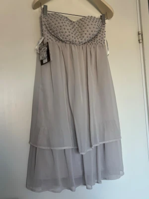 Ljus axelbandslös klänning med pärlor - Snygg grå/beige axelbandslös klänning med pärlprydd överdel och dubbla lager i kjolen för extra volym. Helt oanvänd 