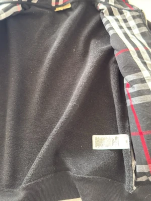 Svart zip hoodie burrbery - Säljer en svart hoodie med dragkedja och huva. Insidan är mjuk och borstad för extra komfort. Ärmarna har rutiga detaljer i svart, vitt och rött som ger en snygg kontrast. Klassisk passform med muddar vid ärmslut och nederkant.