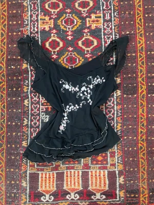 Svart blus med blommig brodyr - Svart blus med v-ringning och volangdetaljer. Blusen har blommig brodyr i vitt och ljusrosa framtill samt spetskantade, korta ärmar. Tunt och lätt material som ger en romantisk känsla.