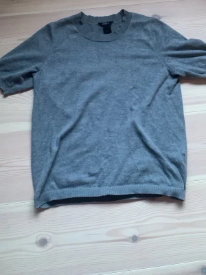 Grå kortärmad topp från Lindex - En enkel och stilren grå topp från Lindex i storlek XS. Toppen har korta ärmar, rund halsringning och är tillverkad i ett mjukt material som känns skönt mot huden. Perfekt att matcha med jeans eller kjol för en clean look.