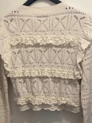 Vit stickad tröja med volanger Zara - Vit stickad tröja från Zara med dekorativa volanger och spetsdetaljer. Tröjan har ett genombrutet mönster, rund halsringning och långa ärmar. Perfekt för dig som gillar romantisk och trendig stil.