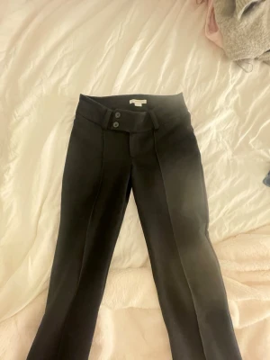 Svarta kostymbyxor med pressveck - Snygga svarta kostymbyxor med låg midja och dubbla knappar i linningen. Byxorna har raka ben och markerade pressveck framtill för en clean look. Perfekta till en dressad outfit och passar till många olika stilar.