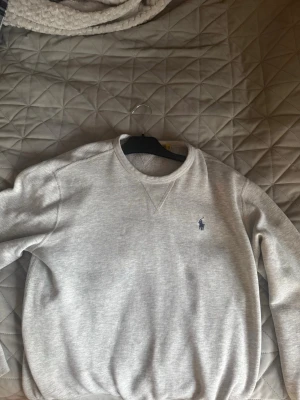 Grå sweatshirt från Polo Ralph Lauren - Klassisk grå sweatshirt från Polo Ralph Lauren med rund halsringning och broderad blå logga på bröstet. Tröjan har ribbade muddar vid ärmslut och nederkant, perfekt för en avslappnad och stilren look.