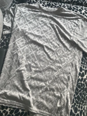 Grå mönstrad t-shirt med logotyp - Snygg grå t-shirt från Calvin Klein med diskret logotypmönster över hela plagget. T-shirten har rund halsringning och korta ärmar. Tillverkad i mjukt och bekvämt material som passar perfekt till en avslappnad stil.
