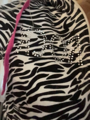 Zebramönstrad hoodie med strass - Snygg hoodie med zebramönster i svart och vitt. Dragkedja framtill, huva med rosa snören och strassdetaljer på bröstet. Muddar i vitt vid ärmslut och midja. Perfekt för dig som vill sticka ut med en cool och trendig stil. Små nästan osynliga fläckar på armen