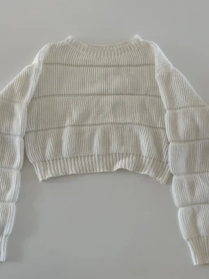 Vit cropped stickad tröja - Snygg vit cropped stickad tröja med breda ärmar och ribbade muddar. Tröjan har ett diskret horisontellt mönster och är perfekt för lager-på-lager-stil. Mjuk och skön, passar till både jeans och kjol. Liten defekt på ärmen som man ser på bilden. Storlek m men passar mig som xs. 