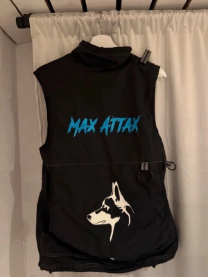 Svart Max Attax väst med hundtryck - Svart väst från Max Attax med blått tryck på ryggen och en vit hundillustration nedtill. Västen har praktiska detaljer som ring för fäste och flera fickor. Perfekt för dig som gillar att sticka ut med en unik look.