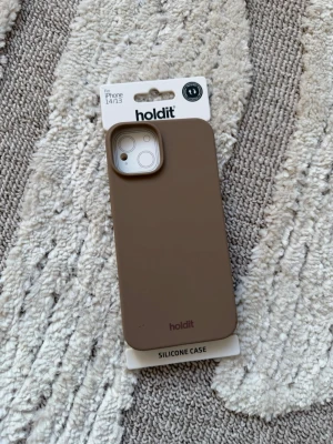 Brunt silikonskal Holdit iPhone 14/13 - Snyggt och stilrent brunt silikonskal från Holdit som passar iPhone 14 och 13. Skalet är gjort i mjukt silikon som ger ett bra grepp och skyddar mobilen mot stötar. Perfekt för dig som vill ha ett minimalistiskt och modernt mobilskal. Helt oanvänd. 
