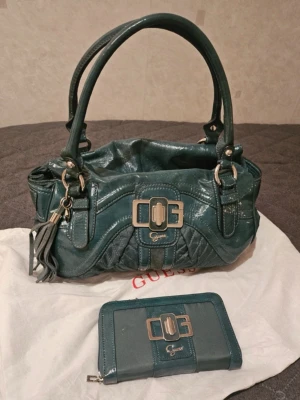 GUESS väska - Turkos/Grön färg på läder, rymlig och superfin modell med gulddetaljer och ikoniskt spänne framtill. Stor nog för laptop, böcker och allt du behöver till jobb/skola. Äkta läder – nypris betydligt högre. Använd, synligt/tydligt slitage finns (se bilder), lädret har gått av på vissa ställen men fortfarande väldigt snygg, därav priset. Säljes då den inte används – för fin för att bara ligga. OBS plånbok ingår EJ  Nypris - runt 3000kr