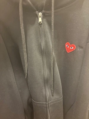Svart hoodie från Comme des Garçons - Svart hoodie från Comme des Garçons Play med dragkedja och huva. Ikoniskt rött hjärta med ögon broderat på bröstet. Mjuk bomullskvalitet och klassisk passform. Perfekt för dig som gillar streetwear och stilrena detaljer.