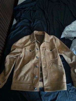 Beige skinnjacka/mocha från Mr P. - Snygg beige mocha jacka från Mr P. i klassisk modell med krage och tryckknappar. Jackan har två bröstfickor med lock och två sidofickor. Tillverkad i mjukt skinn med stilrena sömmar och en avslappnad passform. Perfekt för dig som vill ha en tidlös och trendig look.