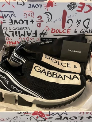 Dolce & Gabbana slip-on sneakers - Svarta slip-on sneakers från Dolce & Gabbana med vit sula och stora logotexter på ovandelen. Skorna har en chunky sula, elastisk ovandel i mesh och vita detaljer längs kanterna. Perfekta för dig som gillar statement-plagg och streetstyle.
