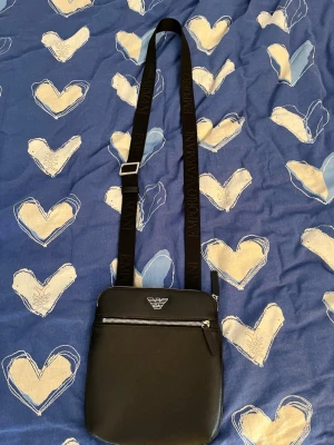 Svart axelväska Emporio Armani - Snygg svart axelväska från Emporio Armani med justerbar axelrem där loggan är invävd. Väskan har en silverfärgad dragkedja framtill och det klassiska Armani-märket på framsidan. Tillverkad i slätt konstläder för en clean look.