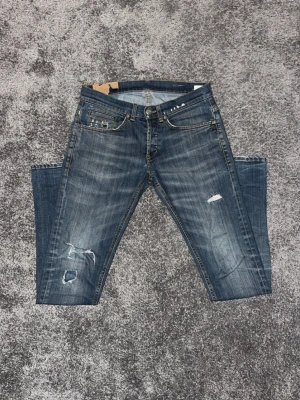 Blå slitna jeans från Dondup med slitningar - Säljer ett par blå jeans från Dondup med slitningar och hål på benen. Jeansen har fem fickor, normal midja och slim-fit passform. Materialet är klassisk denim i bomull och de har en cool, använd look med detaljerade slitningar.