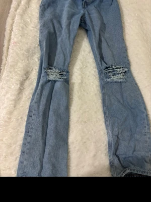 Ljusblå ripped jeans från H&M - Säljer ett par ljusblå jeans från H&M med raka ben och slitna detaljer vid knäna. Jeansen har hög midja och knäppning med fyra silvriga knappar. Perfekta för en avslappnad och trendig look. Använd ett fåtal gånger.😍🤩