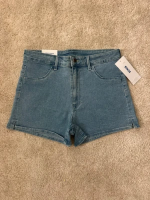 Blå denimshorts från H&M - Klassiska blå denimshorts från H&M med femficksmodell, bälteshällor och knappgylf. Shortsen har en enkel design med raka ben och materialet är stretchigt för extra komfort.