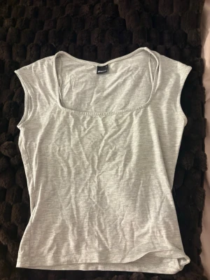 Ljusgrå topp från Gina Tricot - En enkel och stilren ljusgrå topp från Gina Tricot med bred, fyrkantig ringning och ärmlös design. Tillverkad i mjuk bomullsmix som känns skön mot huden. Perfekt att matcha med jeans eller kjol för en clean look.