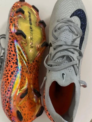 Nike mercurial  - Säljer ett par grå Nike fotbollsskor med svart swoosh på sidan och snörning. Sulan är orange med svarta prickar och har en holografisk finish samt svarta dobbar. Ovandelen är i mesh-liknande material för bra ventilation och passform. Perfekta för dig som vill sticka ut på planen.
