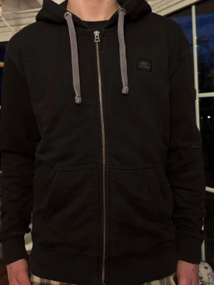 Svart zip-hoodie med gråa snören - Svart hoodie med dragkedja. Har gråa snören i huvan, två stora fickor framtill och ribbade muddar vid ärmslut och nederkant. Klassisk och enkel stil som passar till det mesta. Liten logga på bröstet.