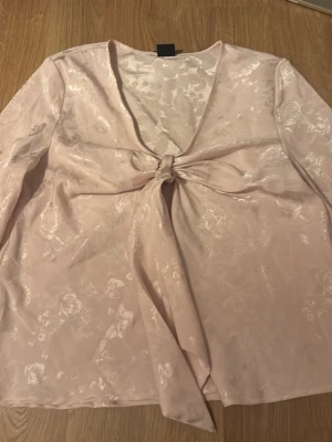Ljusrosa blus med knyt framtill - Snygg ljusrosa blus i glansigt tyg med subtilt blommönster. Blusen har v-ringning och knytning framtill som ger en trendig look. Långärmad modell som passar perfekt till jeans eller kjol. Materialet känns mjukt och har en elegant finish.