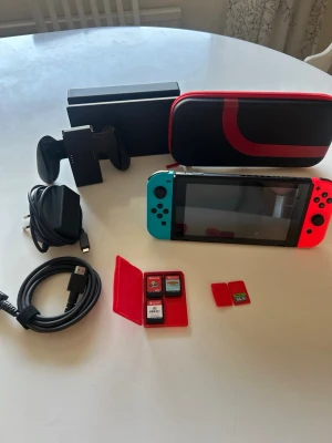 Nintendo Switch med tillbehör och spel - Nintendo Switch med blå och röd Joy-Con, svart dockningsstation, svart och röd bärväska, laddare, HDMI-kabel, handkontrollshållare samt tre spel: Super Mario Odyssey, Donkey Kong Country och FIFA 21. Minneskort på 64GB ingår. Perfekt för gaming hemma eller på språng.