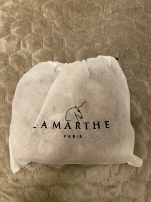 Kamelbrun handväska från Lamarthe - Snygg handväska i kamelbrunt läder från Lamarthe Paris. Väskan har en rundad form, silverfärgat spänne framtill och justerbar axelrem. Perfekt accessoar för att lyfta din outfit med en klassisk touch.