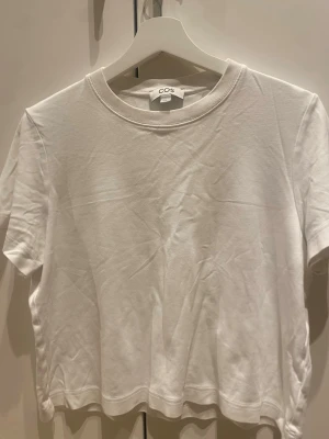 Vit basic t-shirt från COS - En klassisk vit t-shirt från COS i mjuk bomull. Modellen är rak med korta ärmar och rund halsringning. Perfekt till jeans eller kjol för en clean och enkel look.