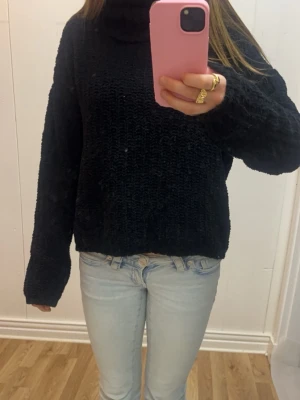 Svart stickad polotröja - Mysig svart stickad polotröja med avslappnad passform och långa ärmar. Tröjan har en hög krage och grovstickad struktur som ger en cool och trendig look. Perfekt att matcha med jeans för en enkel stil.