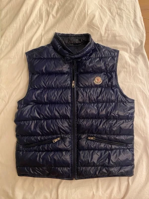 Blå Moncler Gui Väst - Snygg mörkblå dunväst från Moncler med hög krage och dragkedja framtill. Västen har två fickor med dragkedja och det klassiska Moncler-logotypen på bröstet. Tillverkad i glansigt material och fylld med dun för extra värme. Storlek 3 - passar M. Äkta.