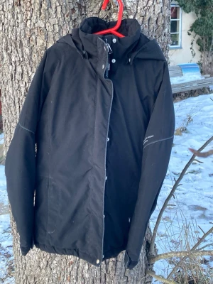 Svart stalljacka med huva och dragkedja - Säljer en svart jacka med huva och dragkedja framtill. Har varit en favorit såväl i stallet som vid ridning kyliga dagar. Jackan har tryckknappar, hög krage och diskreta sömdetaljer. 