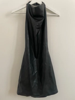Svart halterneck klänning i fakeläder - Snygg svart halterneck klänning i glansigt läderliknande material. Klänningen är ärmlös och har en öppen rygg, vilket ger en trendig look. Perfekt för dig som vill sticka ut med en stilren och modern outfit.