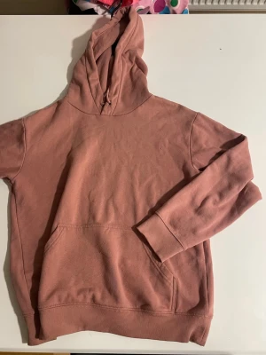 Rosa hoodie med huva och ficka - En enkel och snygg hoodie i dämpad rosa färg. Hoodien har en stor huva med dragsko och en klassisk magficka framtill. Tillverkad i mjukt bomullstyg med ribbade muddar vid ärmslut och nederkant. Perfekt för en avslappnad och chill stil.