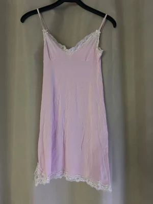 Ljusrosa nattlinne med spetsdetaljer - Sött ljusrosa nattlinne med tunna axelband och v-ringad hals. Dekorerat med vit spets längs urringning och nederkant. Perfekt för mysiga kvällar hemma.