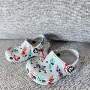 Grå barn Crocs med bilmotiv size 22-23 - Säljer ett par grå Crocs sandaler med färgglada tryck av bilar och fordon. Skorna har klassisk håldesign, justerbar hälrem och är tillverkade i lätt och tålig plast. Perfekta för dig som gillar lekfulla detaljer och bekväma skor.