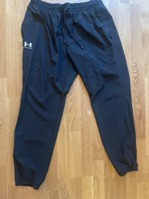 Svarta träningsbyxor Under Armour - Säljer ett par svarta träningsbyxor från Under Armour med resår i midjan och dragsko. Byxorna har logga på vänster ben och är tillverkade i ett lätt och snabbtorkande material, perfekta för gymmet eller löprundan.