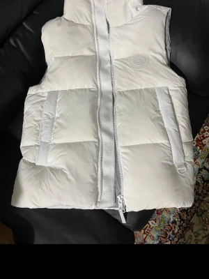 Vit dunväst från Canada Goose - Snygg vit dunväst från Canada Goose med hög krage och två stora fickor framtill. Västen har en diskret logotyp på bröstet i samma färg som tyget och dragkedja framtill. Perfekt för lager på lager och riktigt clean look.