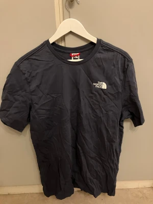 Blå The North Face t-shirt - Mörkblå t-shirt från The North Face med logga på bröstet och stort tryck på ryggen med texten 'Never Stop Exploring'. Klassisk rund hals och korta ärmar. Perfekt för dig som gillar streetwear och outdoor-vibe.