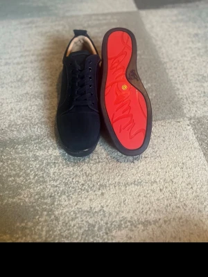 Mörkblåa Louboutins - Snygga svarta sneakers från Christian Louboutin med klassisk röd sula och signaturdetaljer. Skorna har lågt skaft, svarta skosnören och diskret broderad logga på hälen. Ovandelen är i mocka och insidan är ljusbeige.