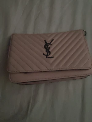 fake-YSL handväska - Säljer en ljusrosa quiltad handväska från YSL med klassiskt YSL-monogram i svart metall på framsidan. Väskan har ett elegant, rektangulärt format och är tillverkad i läder med chevronmönster. Perfekt för dig som vill ha en stilren och lyxig accessoar. Fake. 