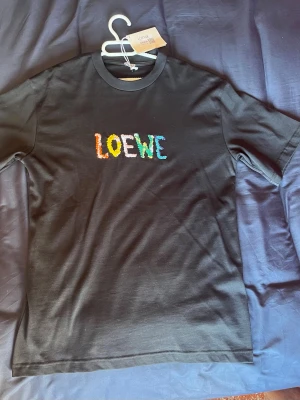 Svart Loewe t-shirt med färgglad logga - Snygg svart t-shirt från Loewe med broderad logga i flera färger på bröstet. Klassisk rund hals och korta ärmar. Tillverkad i mjuk bomull som känns skön mot huden. Perfekt för dig som vill ha en stilren men ändå unik look. Säljs då den aldrig kommit till användning. 